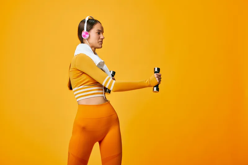 Woman fitness enthusiast in orange athletic outfit holding dumbbells with headphones, performing a stylish workout against a vibrant orange backdrop.