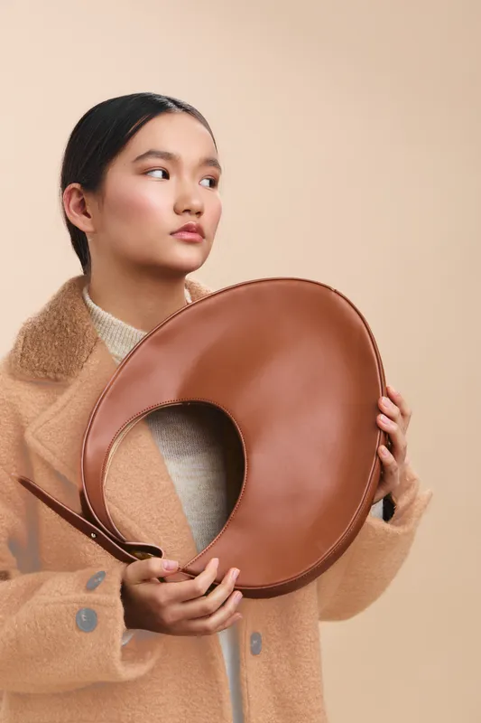 Asian woman gazes to the side while holding a round tan prop, posing in a minimalist studio. The neutral tones and soft lighting create a calm, contemporary mood.