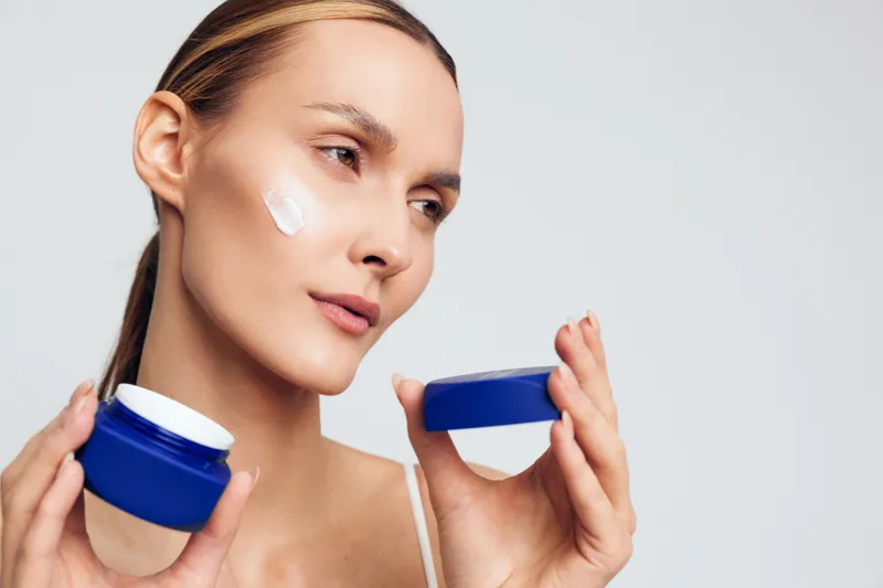 A woman applies moisturizer from a blue jar on her cheek, displaying a skincare routine and a calm self care moment. Focus on hydration, skin health, and everyday beauty.