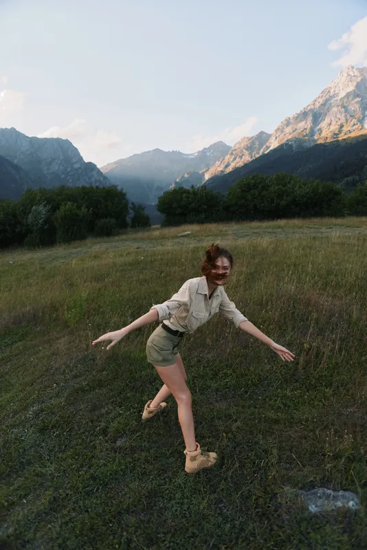 One young woman in a beige shirt and olive shorts balances playfully in a mountain meadow with rugged peaks behind, radiating carefree outdoor energy.