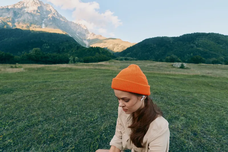 Young woman wearing orange beanie sitting on grass in mountain field during daytime. Concept of lifestyle, nature, and outdoor relaxation with clear sky and scenic landscape.
