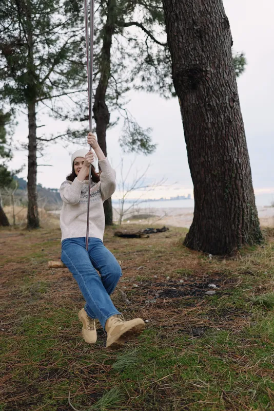 Outdoor swing with a person in casual clothing swinging under tall trees in a park setting showing nature and outdoor relaxation, sunlight filtering through needles, green ground, calm moment,