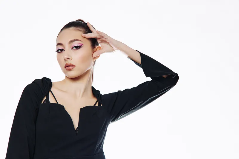 Fashion model with dramatic purple eyeliner poses confidently in a black edgy outfit. White background studio shot puts emphasis on beauty and artistic expression.
