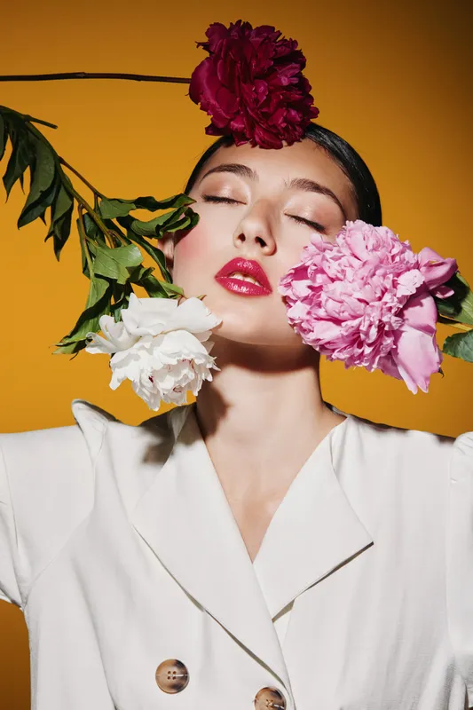 Fashion model with bright glossy lips and flawless skin poses with colorful flowers near face. Creative beauty portrait with dramatic mood on solid yellow background.