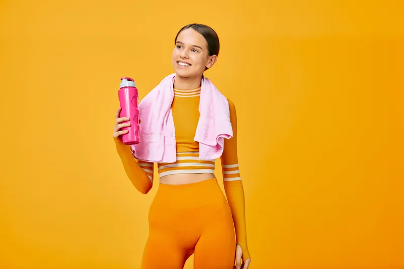 Fitness themed portrait of a smiling woman in bright orange athletic wear, holding a pink water bottle with a towel draped over her shoulders against a bold orange backdrop.