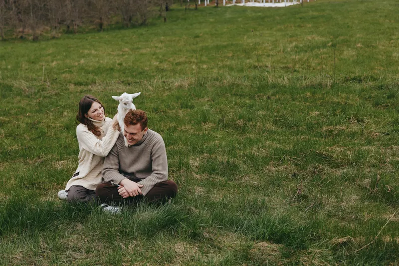 Cheerful couple outdoors, sitting on grass with a playful goat kid between them, sunlit meadow moment, candid interaction, natural colors, relaxed posture, wholesome countryside vibe