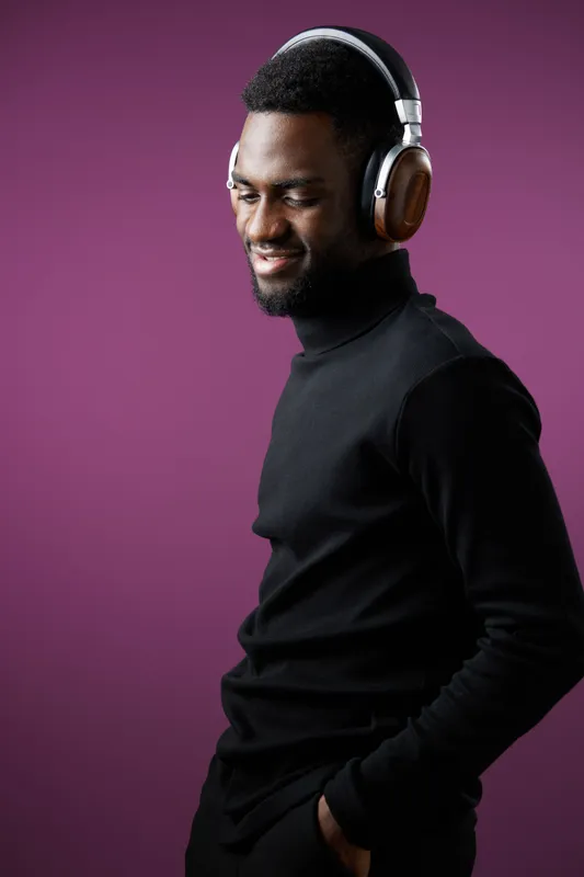 Young African man enjoying music with headphones, wearing a black turtleneck on a vibrant purple background, expressing joy and relaxation through his smile