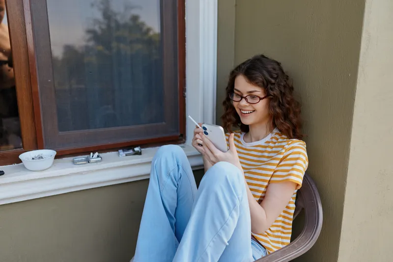 A young woman in a striped yellow t shirt and light blue jeans sits on a balcony railing, smiling softly as she scrolls through her phone in a casual, everyday moment.