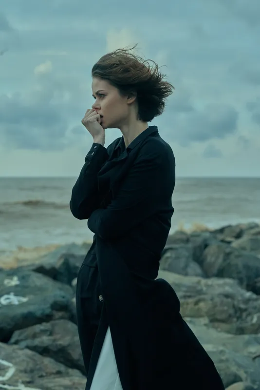Woman in a dark coat on a windy rocky shore, thoughtful gaze toward the horizon, rugged coast and ocean in a moody seascape, solitary mood