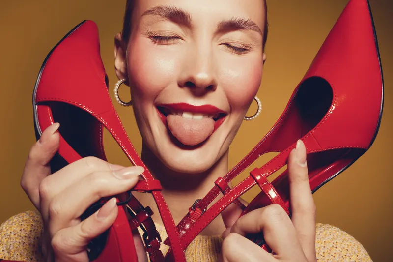 Happy woman with closed eyes holds red high heels near face and shows tongue. Bright studio portrait with warm lighting and yellow background. Concept of fashion, joy, and playful mood.