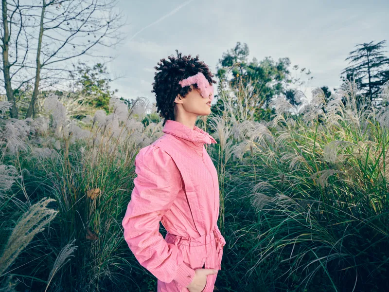 Woman in a pink coat stands in a grassy field, wearing sunglasses, posing for a fashion oriented outdoor portrait with natural light and a calm, airy atmosphere.