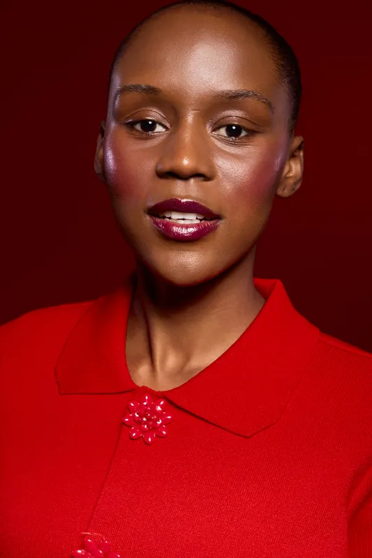 Close-up portrait of a young black woman with dark skin wearing a red top against a red background. The image highlights beauty, fashion, and confident expression in studio lighting.
