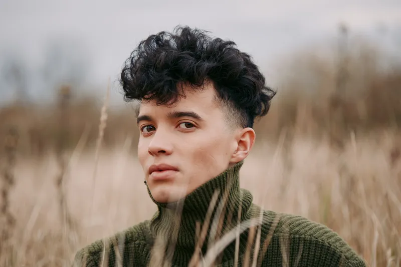 Portrait of a young person with curly hair and a fresh look, wearing a chunky green knit sweater, set against a field of tall grass and a muted sky, conveying authentic presence.