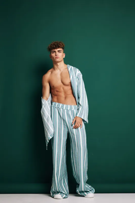 Young man posing shirtless in striped pajama pants and unmatched striped shirt against dark green background, fashion photo with casual and relaxed style studio portrait.