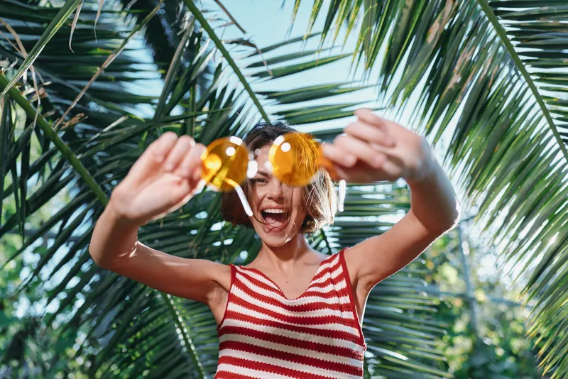 An energetic tropical scene features a joyful woman in a striped top playfully posing with sunglasses, smiling broadly under palm leaves, exuding carefree summer vibes.