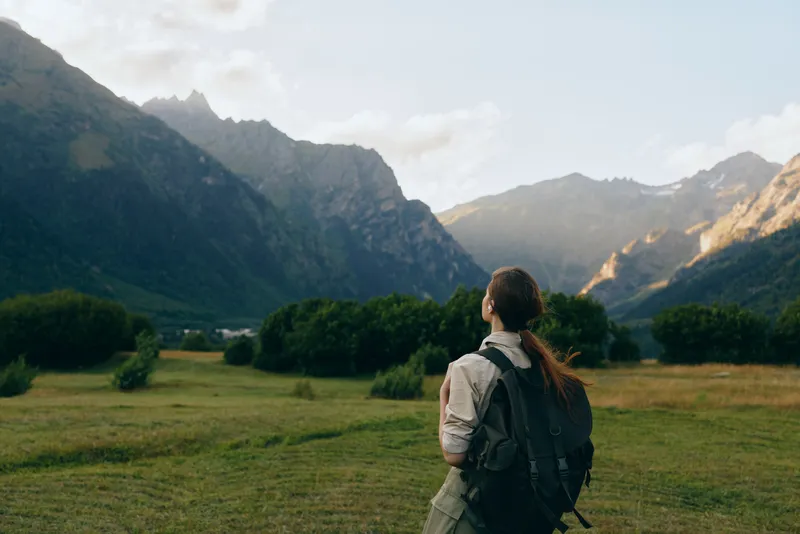 Young woman with a backpack treks through a mountain valley, gazing ahead with a sense of adventure and calm amidst towering peaks, open skies, and lush fields