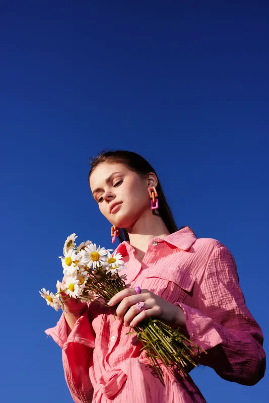 Young woman in a pink shirt holding daisies against a bright blue sky, showcasing a joyful expression and vibrant summer vibes