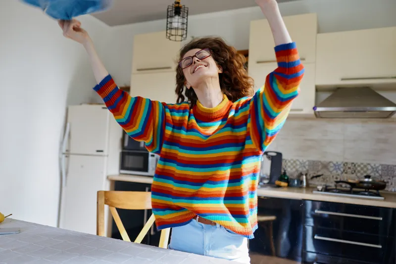 Woman lifestyle at home, energetic moment in kitchen, colorful striped sweater, playful pose as she tosses a hat, bright natural light, candid joyful mood, indoors scene.