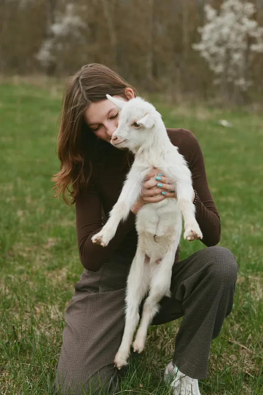 A woman gently holds a young goat in a grassy meadow, capturing a tender lifestyle moment of affection and care between human and animal in a natural rural setting.