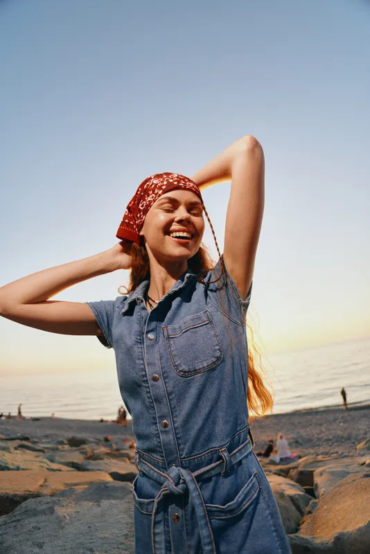 Lifestyle woman in modern boho-western denim street style smiling joyfully at sunset beach with film color effect, enjoying freedom and relaxed mood outdoors.