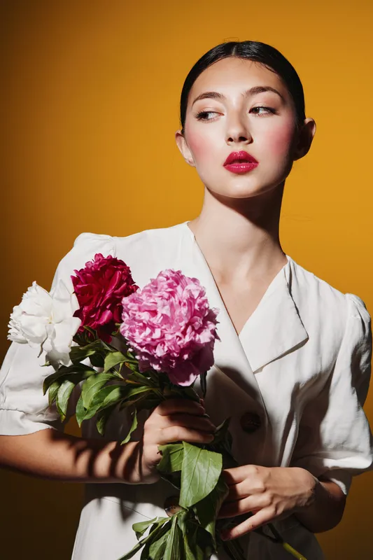 Young woman with flawless makeup and glossy lips holds fragrant peonies in vibrant pink, red, and white. Styled hair and white jacket contrast with warm yellow background. Beauty fashion creative