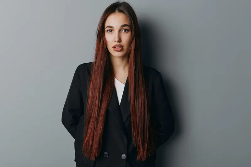 Young woman with long straight hair wearing black blazer standing against gray background looking at camera with neutral expression. Portrait of confident female model with natural makeup.
