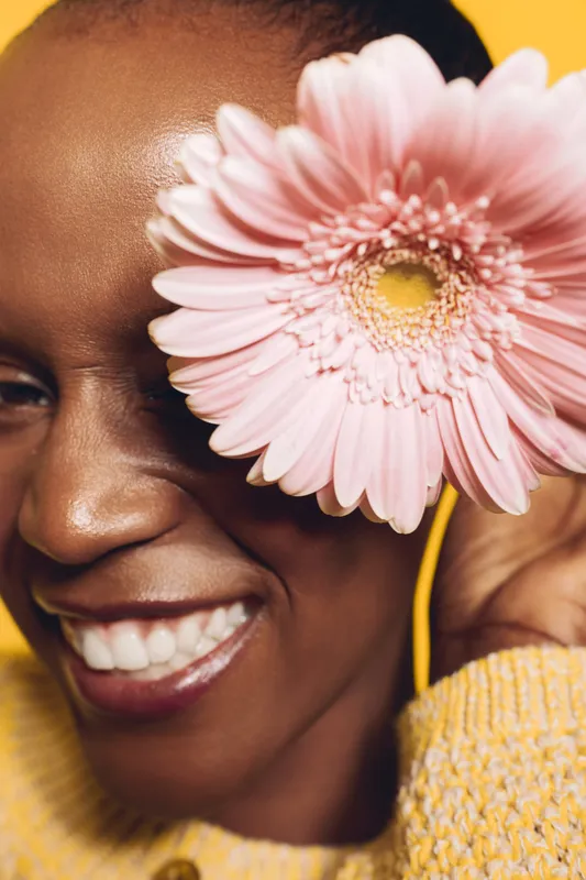 Close-up of smiling African woman with dark skin holding pink flower near eye against yellow background. Concept of happiness, beauty, natural skincare, and positive emotions in studio.