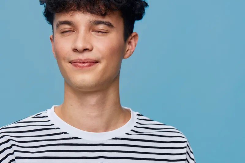 Young man with closed eyes and relaxed smile wearing a striped shirt against a solid blue background expressing calm and peace in a casual style.