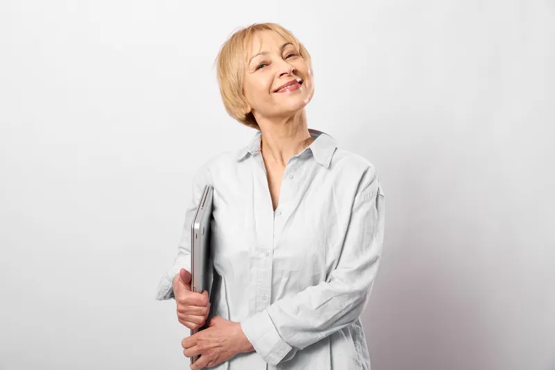 Elderly woman smiling, holding pen for writing, friendly portrait in studio light, casual shirt, calm expression, neutral white background, soft shadows today