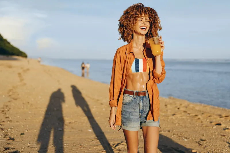 Young woman with curly hair laughs on a sunny beach, holding a juice glass while wearing an orange shirt over a striped bikini and denim shorts, with a calm sea in the background.