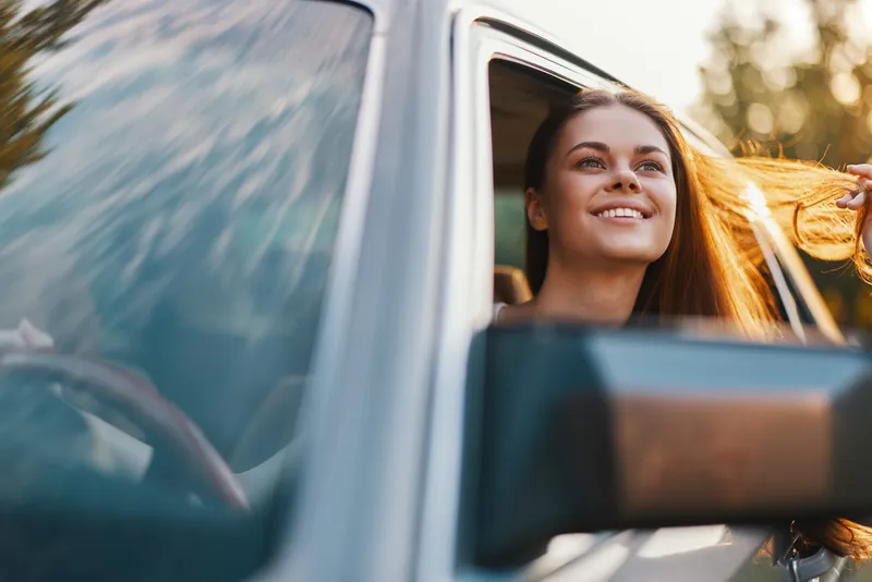Happy young woman enjoying a road trip with her hair flowing in the wind, seated in a car, surrounded by nature's beauty on a sunny day.