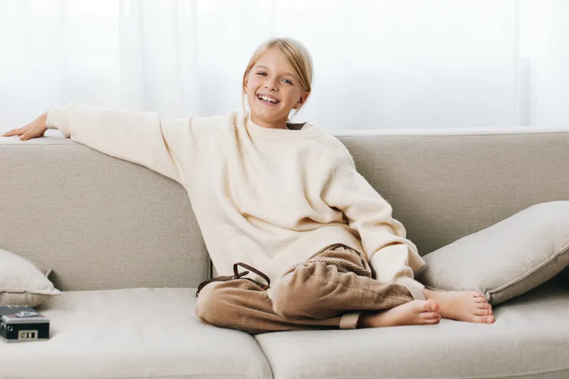 A cheerful child lounges on a light sofa, relaxed and smiling, wearing a soft sweater and casual pants, creating a warm, cozy scene of home comfort and carefree ease.
