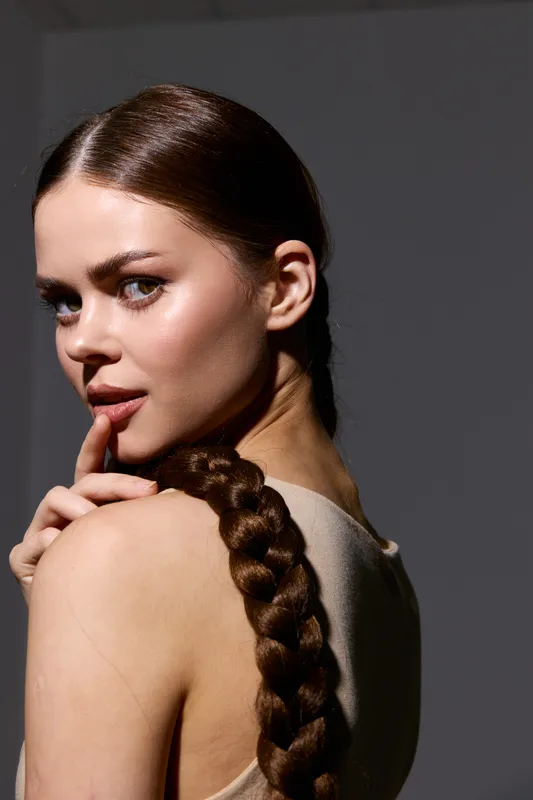 Fashion woman with long braided hair posing thoughtfully against dark background, showcasing natural makeup, smooth skin, and minimalistic style in soft lighting.
