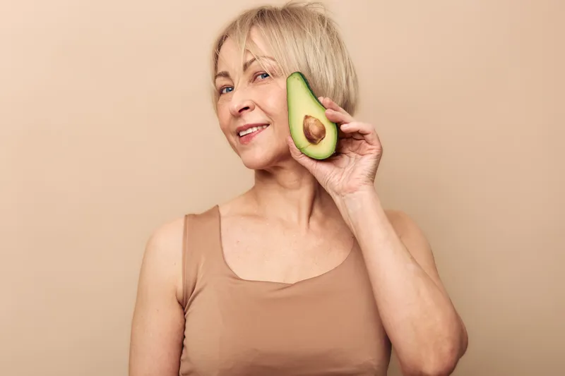 Mature woman with short blonde hair holding half of fresh avocado near face smiles softly. Beige background and neutral clothing create natural skincare and healthy lifestyle concept.