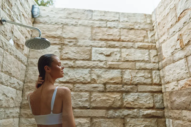 Woman taking a refreshing outdoor shower under rain showerhead against textured natural stone wall on a sunny day, enjoying relaxation and self-care moments.