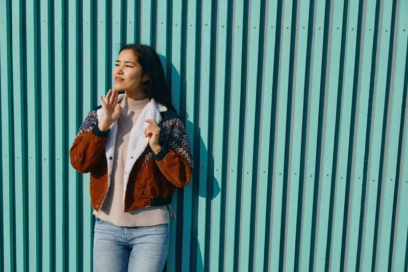 Woman wearing a textured brown jacket and light sweater poses against a blue striped wall, exuding casual street fashion, relaxed confidence, and urban style.