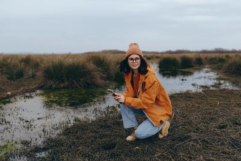 Woman lifestyle portrait outdoors in nature, kneeling by field water, wearing orange jacket and knit hat, smiling softly in autumn landscape near reeds, gentle daylight.