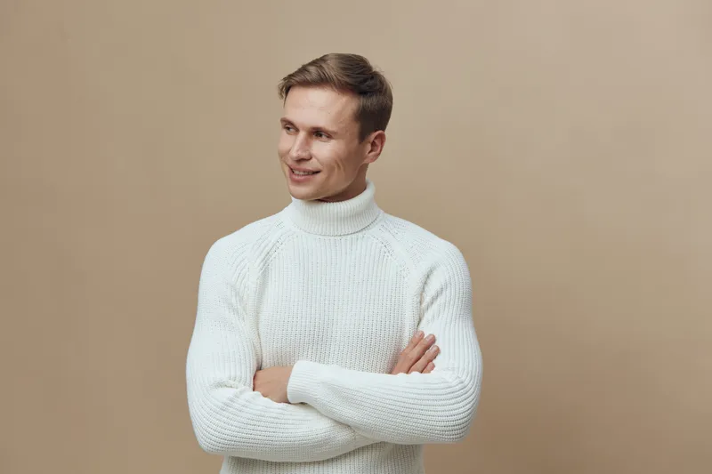 Smiling man in white sweater look away cross arms studio portrait casual wear beige background mental health gadget concept
