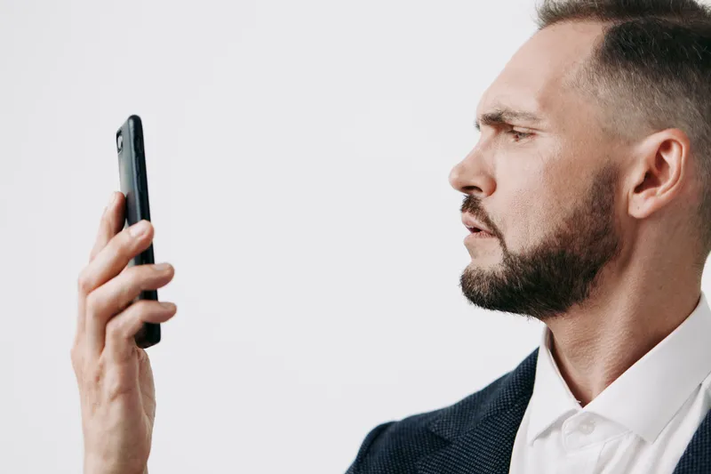Portrait of a professional businessman, focused and confident, examining a smartphone. The subject stands against an isolated colored backdrop, conveying determination and engaged emotion in a modern