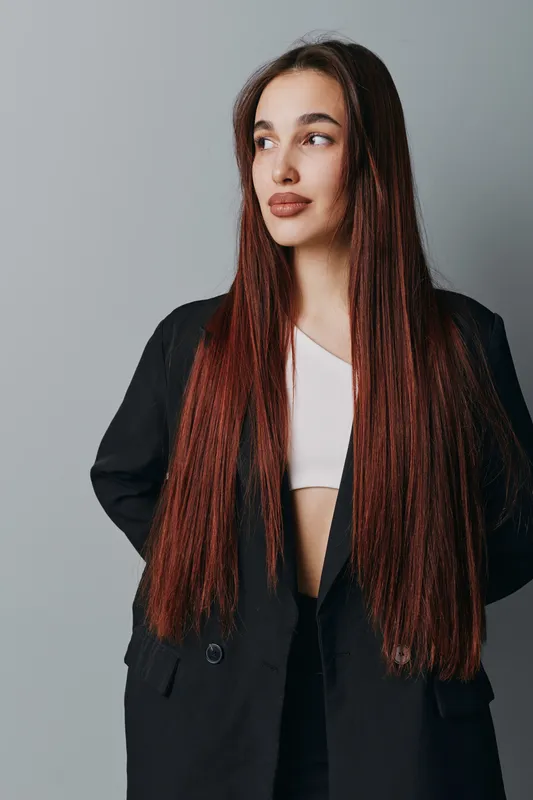 Young woman with long straight hair wearing black blazer and white top standing against gray background looking aside with confident expression and natural makeup in studio.