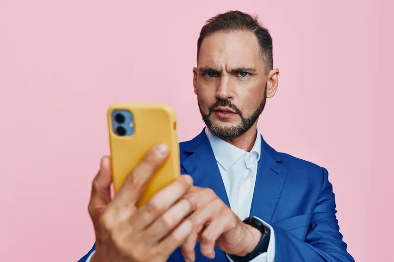 A focused business professional stands against a soft pink backdrop, examining a smartphone with a serious, thoughtful expression, conveying concentration, digital communication, and contemporary