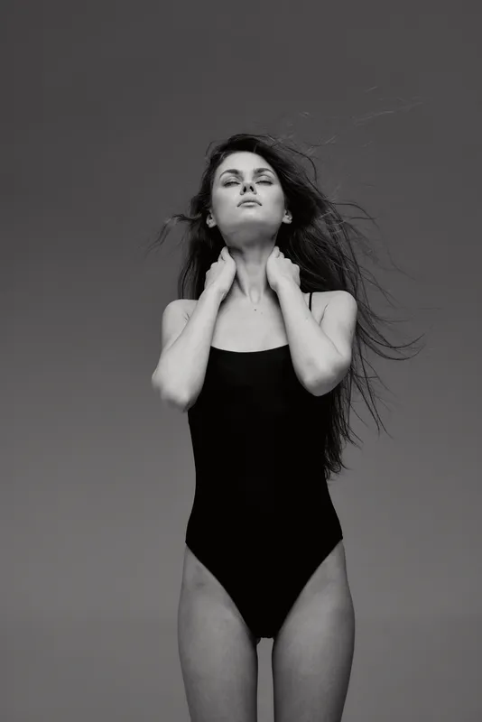 Black and white portrait of a confident woman in a black swimsuit with tousled hair, posing gracefully against a grey background.