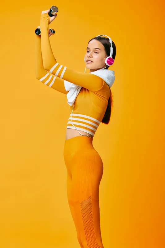 Female fitness model in orange activewear lifting dumbbells, wearing wireless headphones and towel draped over shoulders against a solid vibrant orange studio background, sport concept.