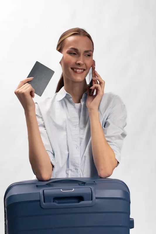 Smiling woman holding boarding pass talks on phone near blue suitcase. Travel concept with happy traveler ready for vacation or business trip, isolated on white background.