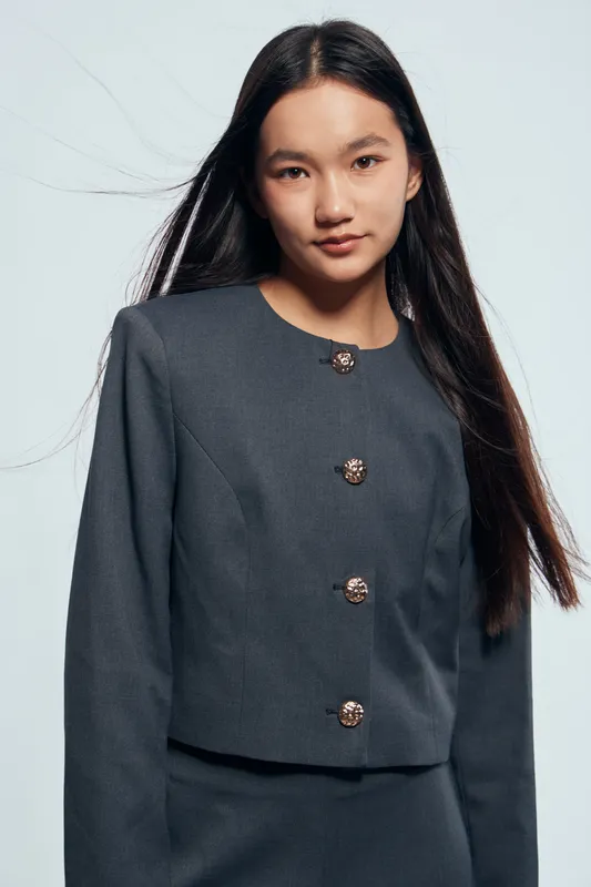 Young woman with long hair wearing a dark gray suit jacket with buttons, posing confidently against a light background in a studio portrait. Minimalist fashion and elegance.