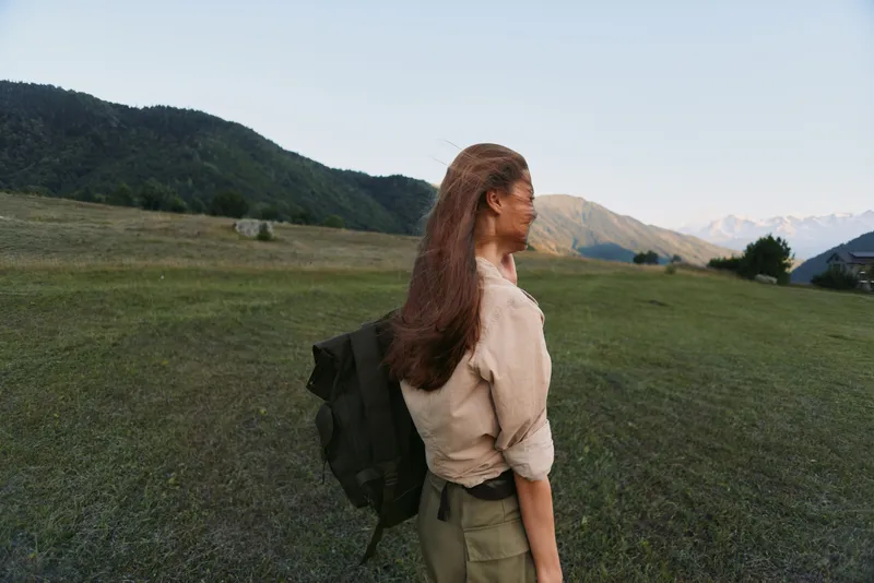 Young woman with long hair stands in open field wearing casual clothes and backpack enjoying nature and mountain landscape during outdoor lifestyle activity.