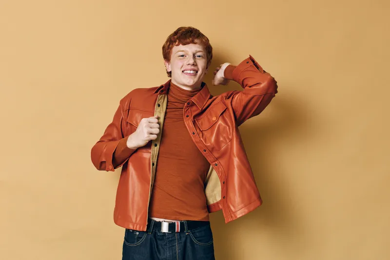 Young man with red hair wearing orange jacket and turtleneck poses smiling against beige background indoors casual style fashion portrait.
