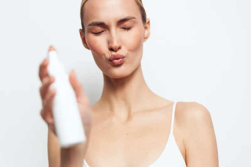 A young woman with clear skin applies facial mist spray with closed eyes and puckered lips. She wears a white tank top against a plain white background. Concept for skincare and beauty.