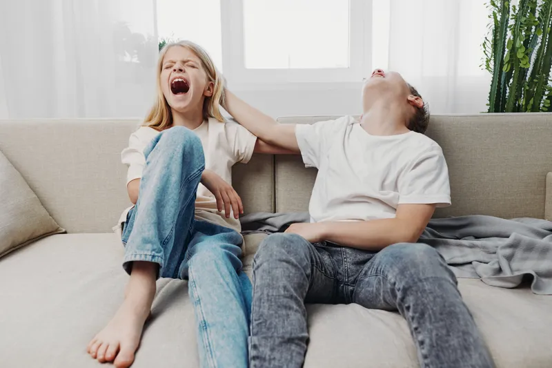 Two children laughing loudly while sitting on sofa in casual clothes in bright living room, having fun and playful moment together.