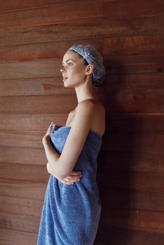 woman in towel wearing shower cap standing against wooden wall, serene expression, natural light highlighting calm and relaxed mood in spa setting